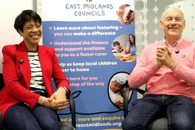 Valerie and Austin sit smiling in front of an informational banner at a Foster for East Midlands event